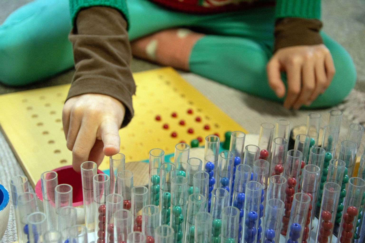 Des mains d’enfant prennent des perles de couleur rangées dans des tubes, devant une planche de calcul Montessori