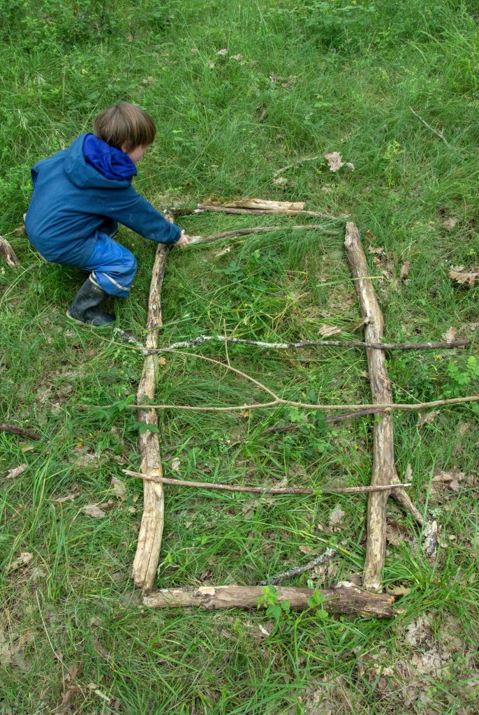 Dans l’herbe, un enfant en combinaison de pluie, vu de dos, assemble des branches pour une œuvre de land art