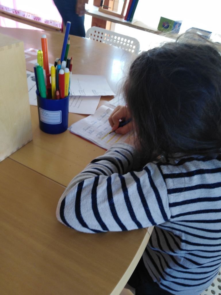 Une élève, vue de dos, écrit dans son cahier, à côté d’un pot de crayons de couleur