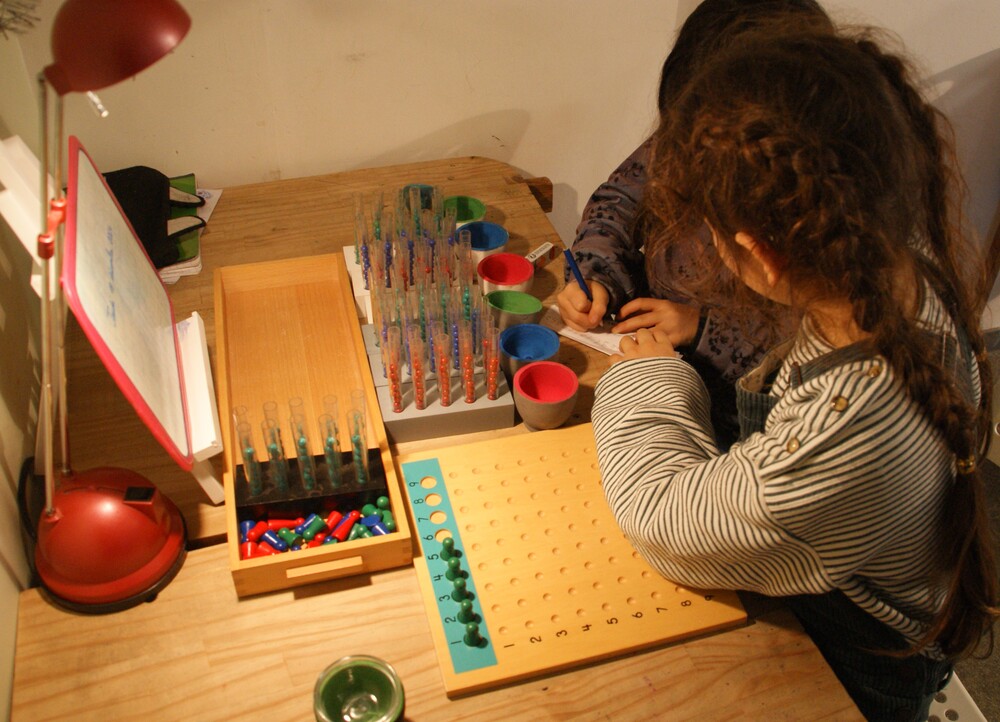 Deux élèves, vus de dos, travaillent à une table avec du matériel de calcul Montessori : tubes de perles, coupelles de couleur et planche à chevilles