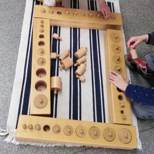 Sur un tapis rayé, des mains d’enfant replacent des cylindres en bois dans les blocs d’encastrement du matériel Montessori
